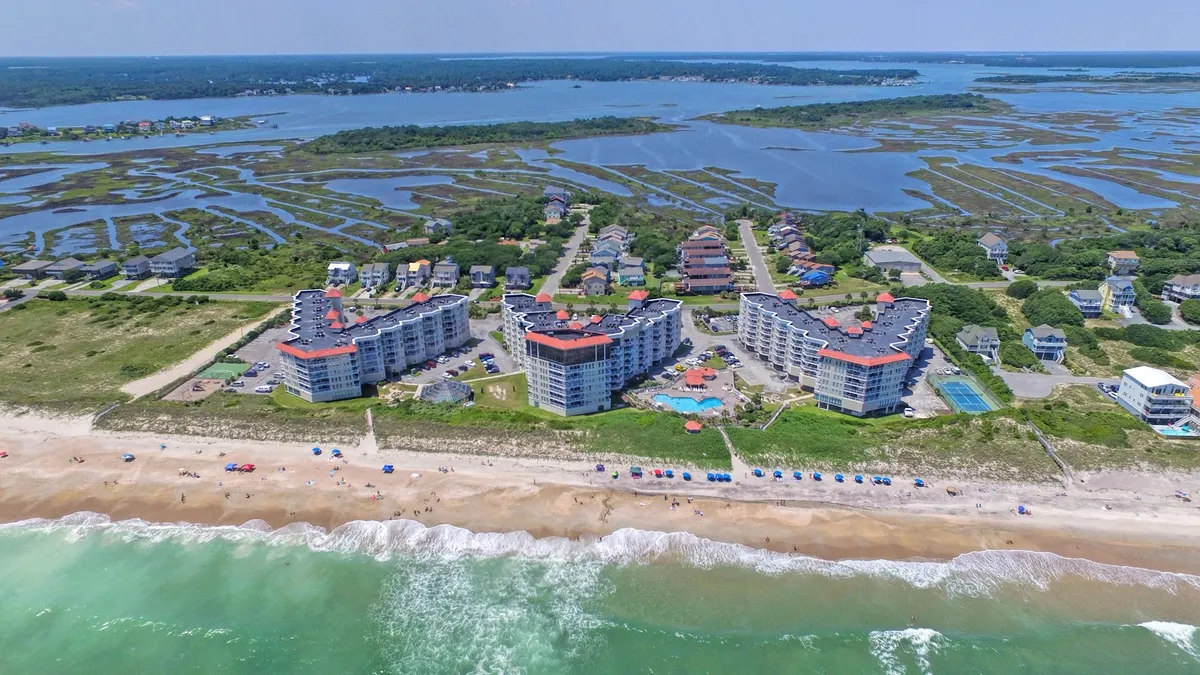 An aerial perspective captures the beachfront condominium property, showcasing the swimming pool, expansive blue ocean, and adjacent marshland. The scene is lively with opportunities for swimming, sunbathing, and exploration.