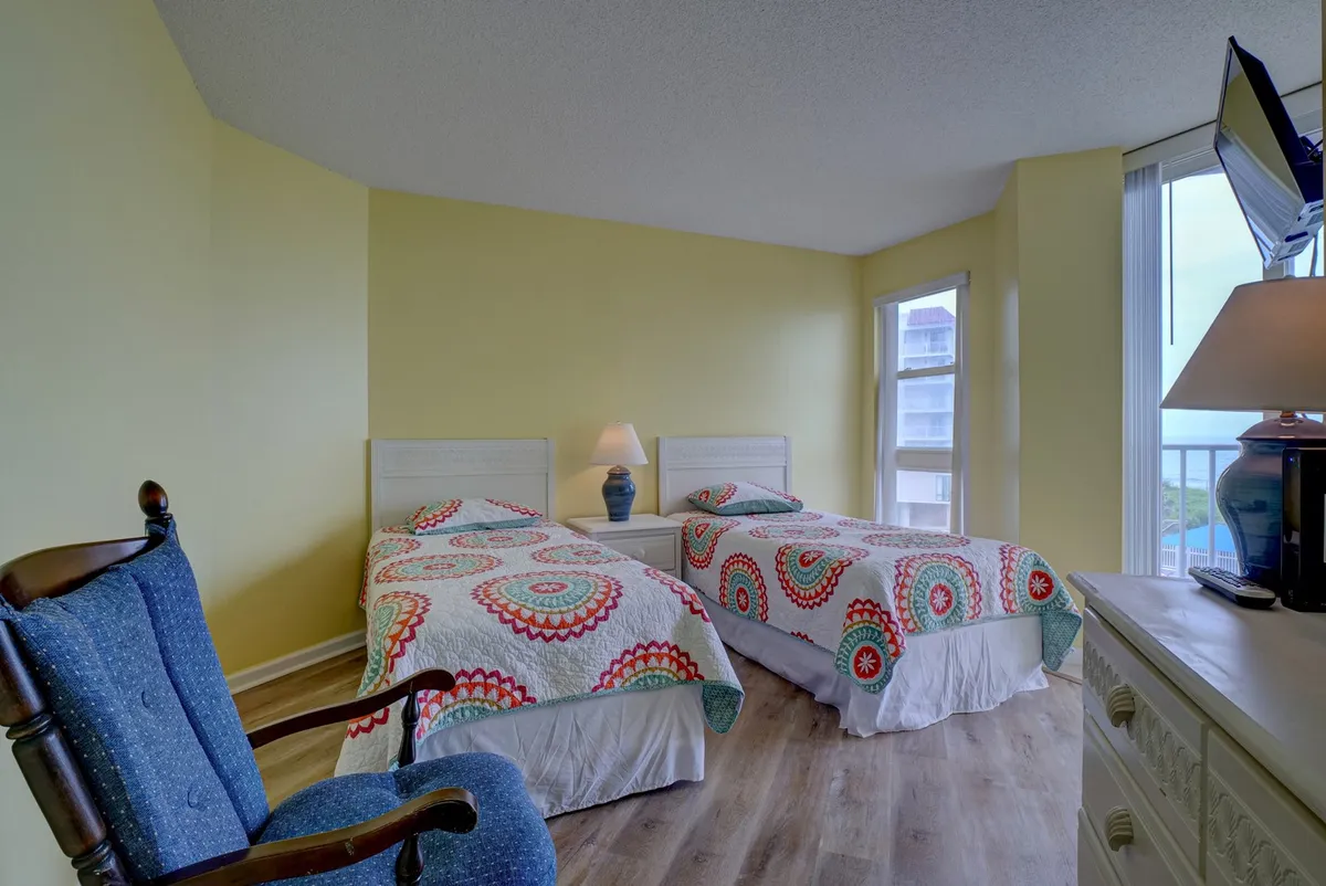 A cheerful second bedroom with two twin beds covered in colorful bedding, a cozy rocking chair, a lamp on the bedside table, and a television on the dresser, all set in a bright, inviting space suitable for kids or friends.