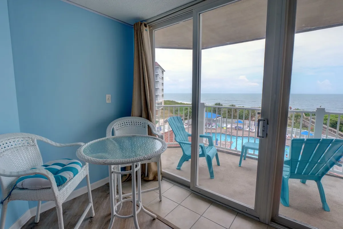 An inviting sunroom or enclosed patio featuring a glass table and white wicker chairs, set against a tranquil blue wall. Through the windows, the ocean is visible, making this a perfect space for sipping coffee, reading, or watching the sunrise.