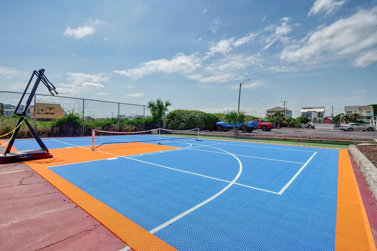 An exterior basketball court equipped with a tennis net, surrounded by greenery and under a clear blue sky. The space is designed for both basketball and tennis or pickleball, encouraging friendly competition and community events. The active mood supports group recreation and social interaction.