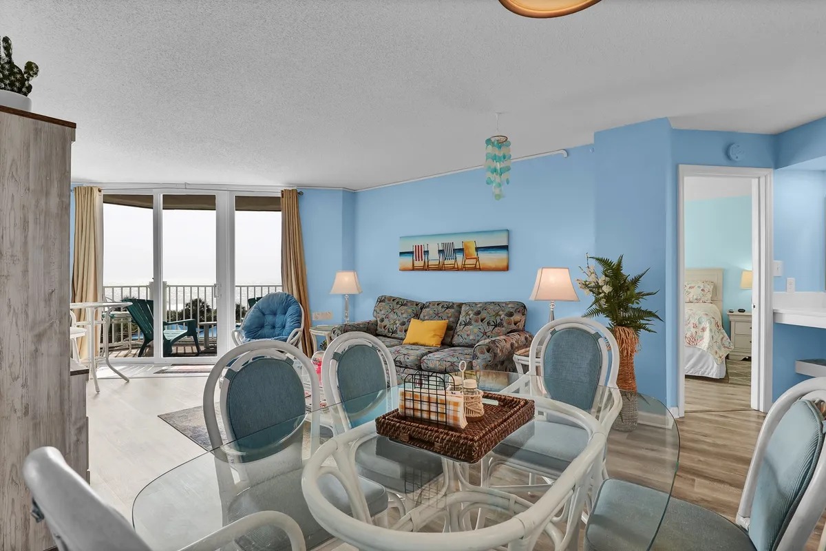 This image features a bright, welcoming open-plan living and dining area, with a colorful couch, large window offering a sweeping ocean view, and a dining table adorned with a decorative centerpiece. Natural light floods the space, creating an inviting setting for both relaxation and shared meals.