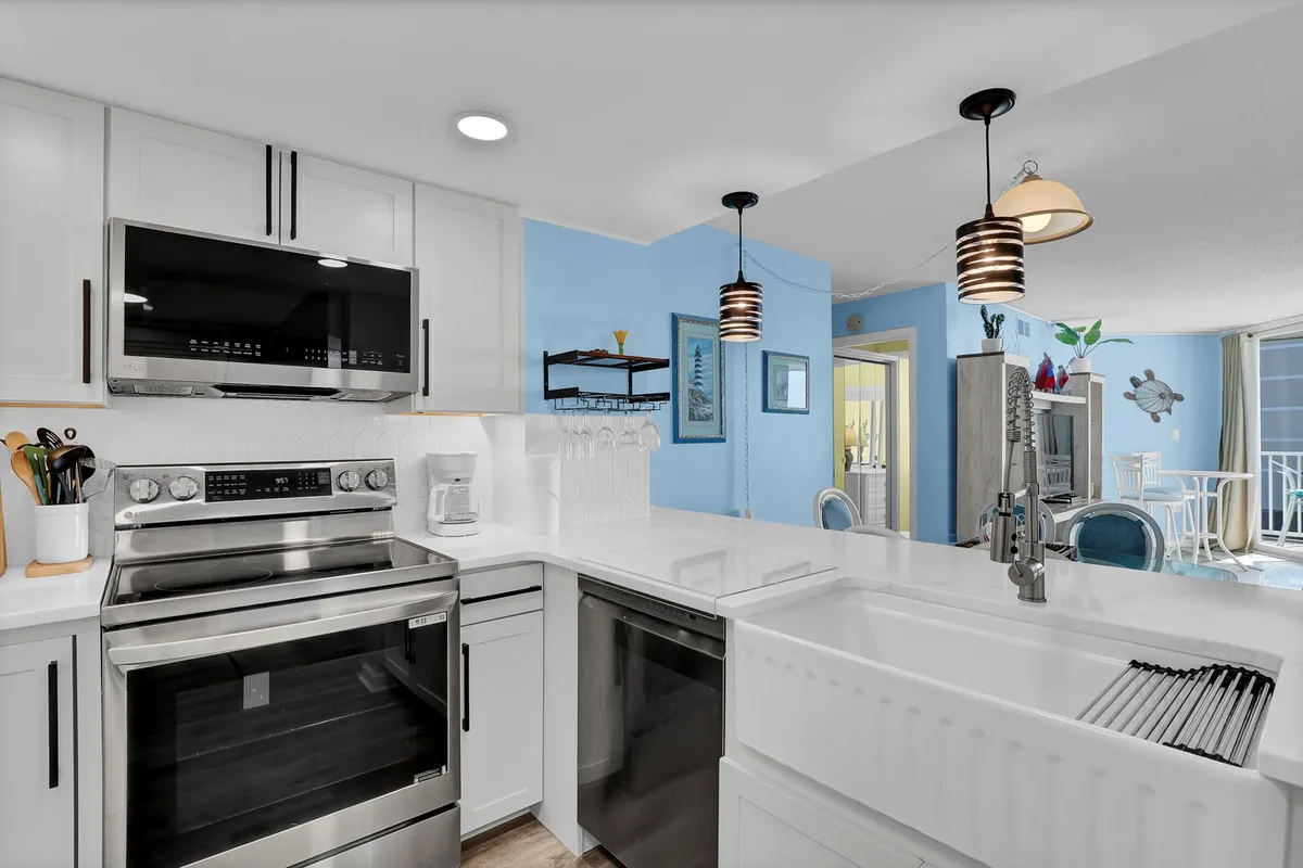 This image features a bright, modern kitchen with stainless steel appliances, a white farmhouse sink, a bold blue accent wall, and contemporary hanging light fixtures. The space is lively and functional, designed for both cooking and entertaining, with an inviting atmosphere perfect for culinary activities.