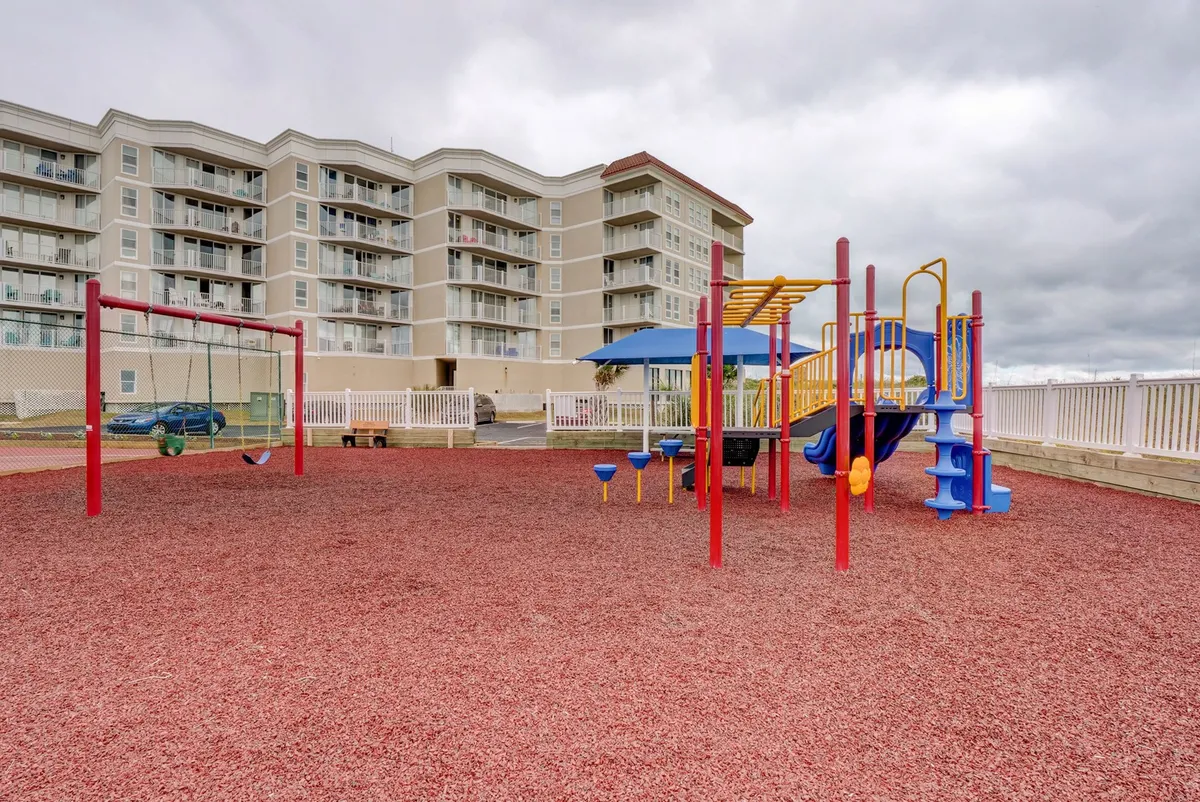 A vibrant playground featuring colorful swings and other play equipment, located near a multifamily building. Children can be seen playing and enjoying themselves, while families gather on the surrounding grassy areas. The overall mood is joyful and energetic, emphasizing family-friendly fun and outdoor recreation.