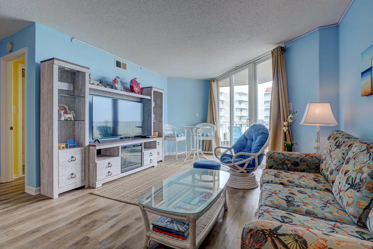 A cheerful living room with a colorful sofa, glass coffee table, and a television unit, all oriented toward broad windows that frame ocean vistas. The space suggests comfort and togetherness, with ample room for family activities or quiet relaxation.