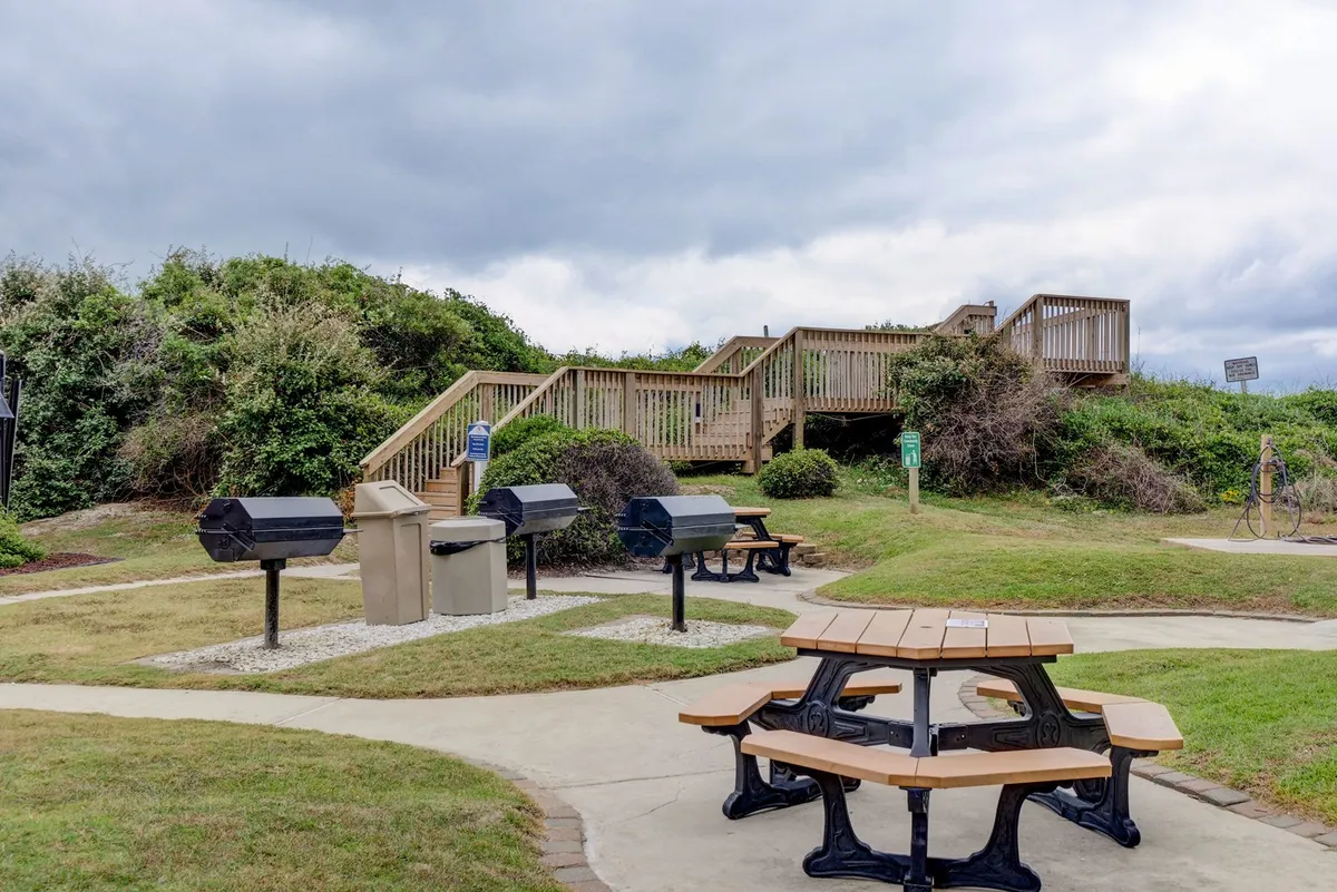 This image features a communal picnic area with barbecue grills, a picnic table, and surrounding greenery. The space is designed for outdoor meals, gatherings, and enjoying the natural environment.