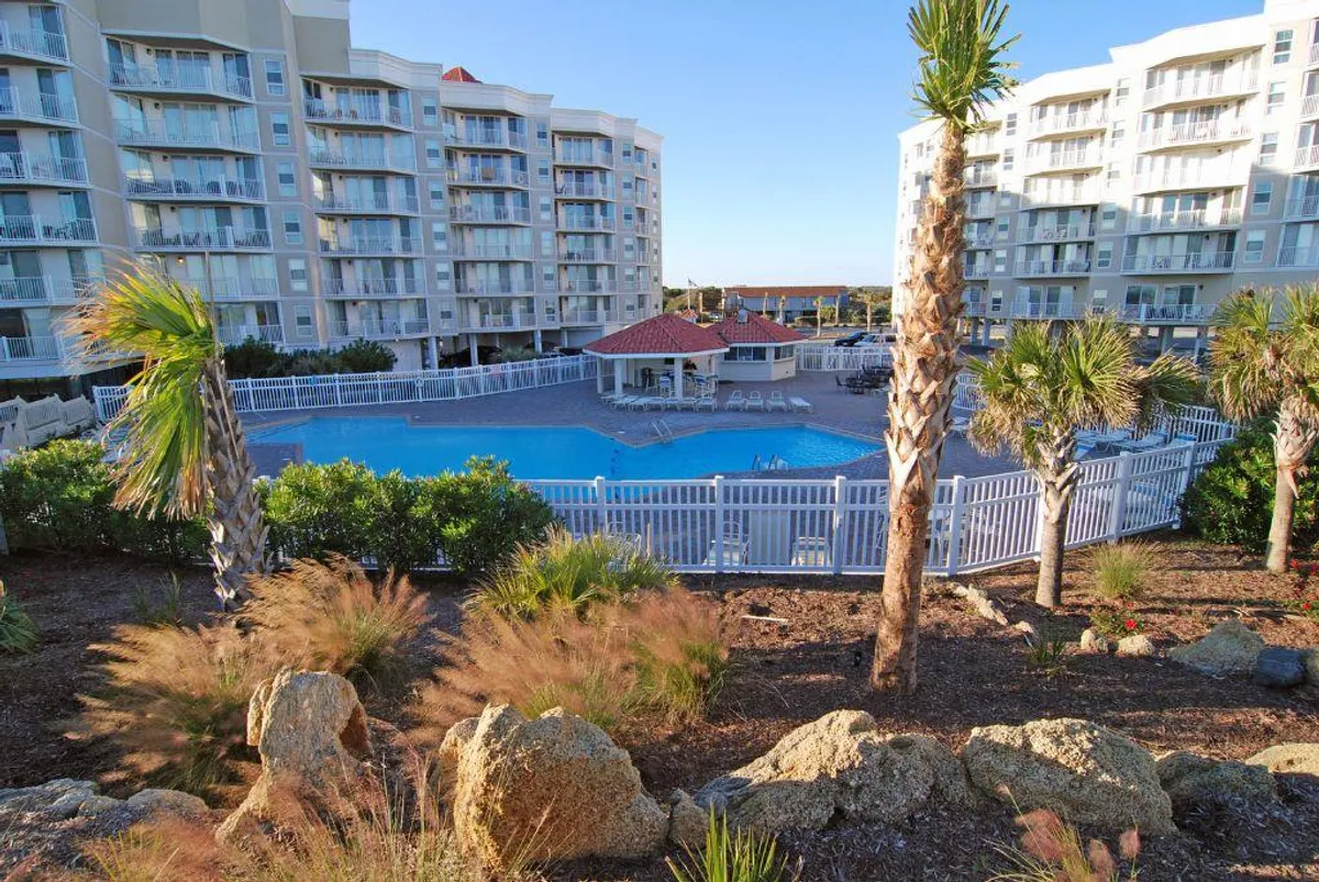 A spacious outdoor swimming pool surrounded by palm trees, with lounge chairs and umbrellas set up around the area. The pool is positioned adjacent to the building's facade, offering an inviting setting for swimming, sunbathing, and social gatherings. The mood is tranquil and relaxing, ideal for families or friends to enjoy resort-style amenities.