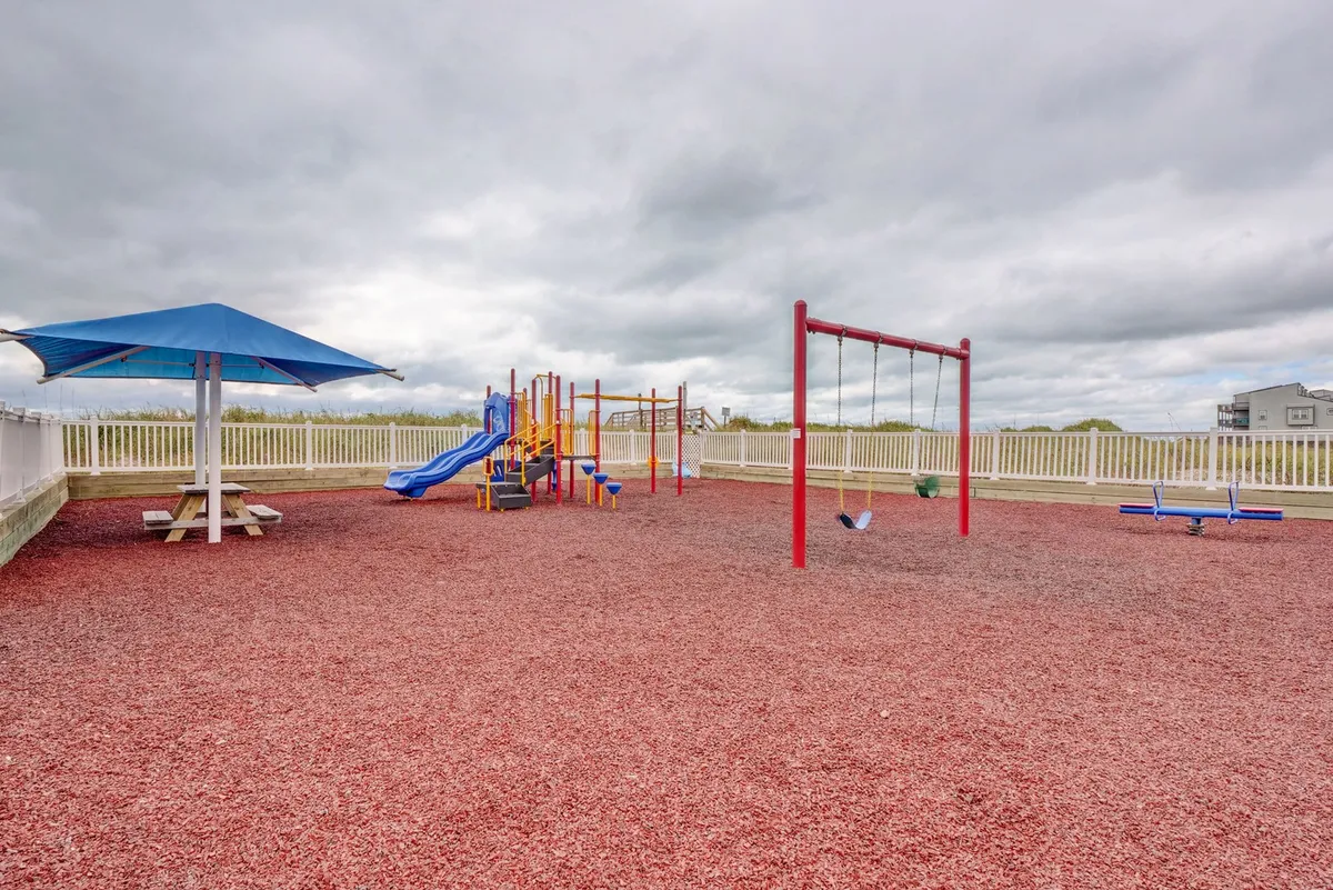A vibrant playground featuring swings, climbing structures, and a picnic table sits beneath a shade structure, inviting children and families to play and relax. The joyful atmosphere and family-oriented design highlight the resort's appeal for guests with kids.