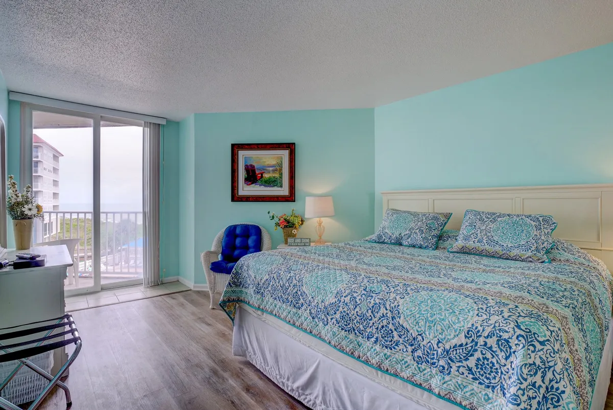 A spacious master bedroom featuring a large bed with patterned bedding, a blue accent chair by the window, framed artwork, and sunlight streaming in through a window, creating a tranquil and comfortable ambiance.